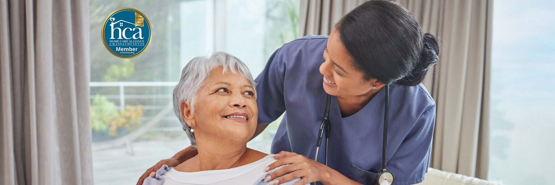 Nurse in scrubs caring for a smiling elderly woman, with a Home Care Alliance of Massachusetts badge visible on the left side.